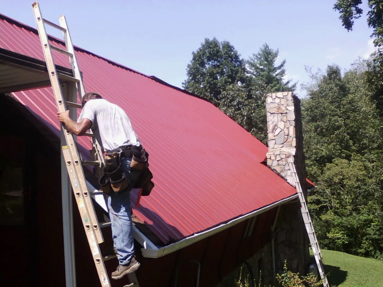Roofer on a ladder beside a red metal roof and stone chimney — Soffit & Fascia Repair by Roofing Brothers in Follansbee